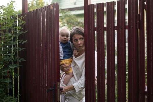 Curious woman with a baby peeps openint the gate of fence, she looks with interest wwhat her neighbor is doing