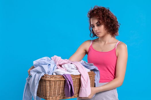 A woman with an angry face carries a wicker basket with shirts for washing. Daily household chores. Young attractive girl with curly hair.