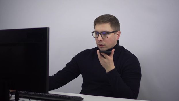 A young IT specialist records a voice message on a smartphone. A man sits in the office at his workplace at the computer. A man with glasses and a black turtleneck. 4k