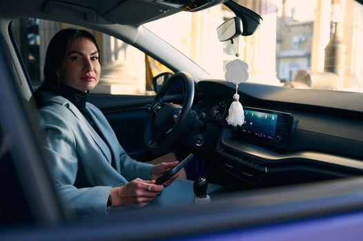 Middle-aged positive pleased confident business woman, executive director, female boss in formal wear, sitting in a car and using digital tablet, telecommuting, planning meetings, smiling at camera