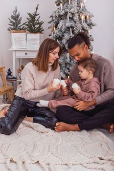 A young diverse family with a baby boy is sitting on a knitted blanket near a Christmas tree, in a room, holding large candles in their hands. Vertical