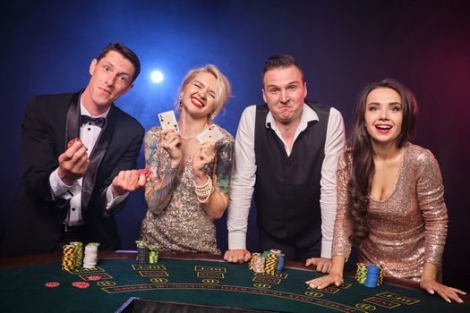 Group of a happy rich companions are playing poker at casino. Youth are making bets waiting for a big win. They are smiling standing at the table against a red and blue backlights on black background. Risky gambling entertainment.