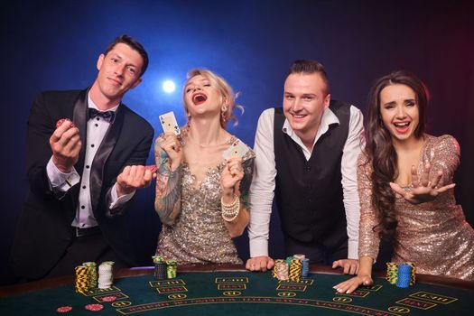 Group of a happy rich colleagues are playing poker at casino. Youth are making bets waiting for a big win. They are standing at the table against a red and blue backlights on black background. Risky gambling entertainment.