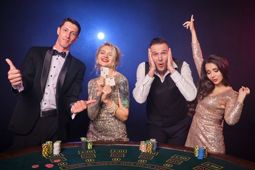 Group of a happy rich friends are playing poker at casino. Youth are making bets waiting for a big win. They are looking overjoyed standing at the table against a red and blue backlights on black background. Risky gambling entertainment.