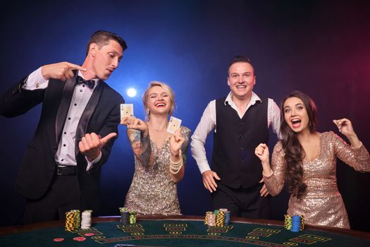 Group of an elegant rich companions are playing poker at casino. Youth are making bets waiting for a big win. They are looking joyful standing at the table against a red and blue backlights on black background. Risky gambling entertainment.