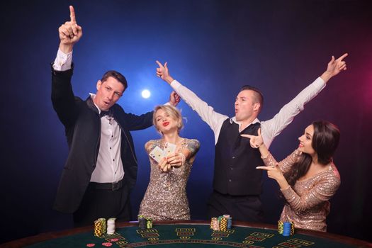 Group of an elegant rich classmates are playing poker at casino. Youth are making bets waiting for a big win. They are celebrating their victory standing at the table against a red and blue backlights on black background. Risky gambling entertainment.