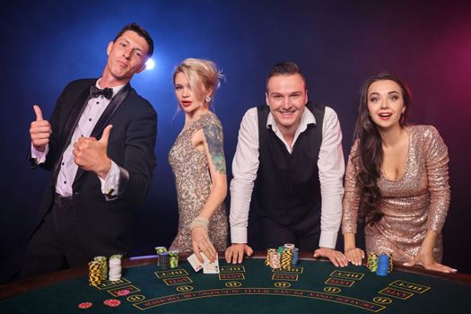 Group of a stylish rich classmates are playing poker at casino. Youth are making bets waiting for a big win. They are standing at the table against a red and blue backlights on black background. Risky gambling entertainment.