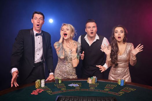 Group of a stylish rich friends are playing poker at casino. Youth are making bets waiting for a big win. They are standing at the table against a red and blue backlights on black background. Risky gambling entertainment.