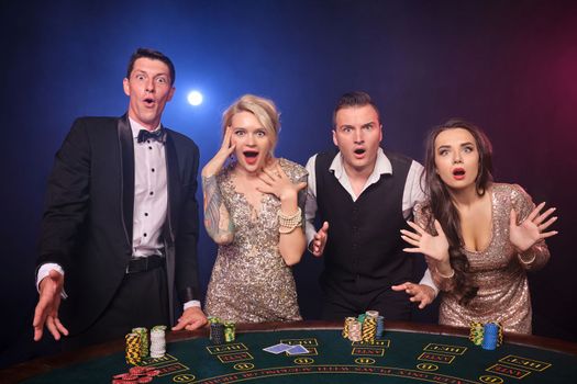 Group of a wondered rich buddies are playing poker at casino. Youth are making bets waiting for a big win. They are looking at the camera standing at the table against a red and blue backlights on black background. Risky gambling entertainment.