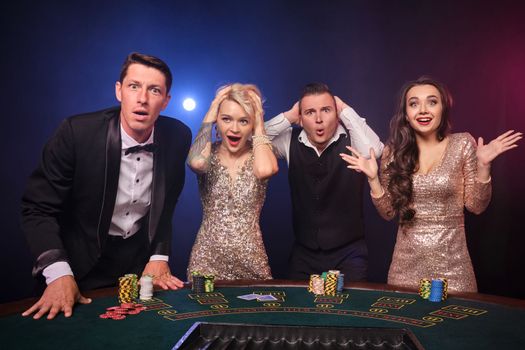 Group of a wondered rich colleagues are playing poker at casino. Youth are making bets waiting for a big win. They are standing at the table against a red and blue backlights on black background. Risky gambling entertainment.