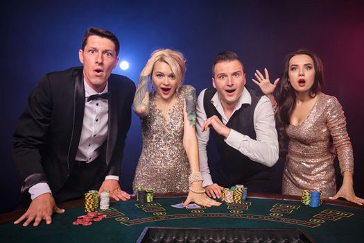 Group of a wondered rich classmates are playing poker at casino. Youth are making bets waiting for a big win. They are looking amazed standing at the table against a red and blue backlights on black background. Risky gambling entertainment.