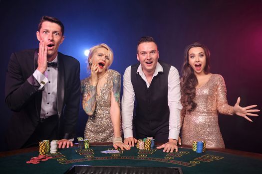 Group of a rich friends are playing poker at casino. Youth are making bets waiting for a big win. They are standing at the table against a red and blue backlights on black background. Risky gambling entertainment.