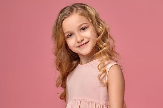 Sweet little girl with a blond curly hair, in a pink dress poses fot the camera and looks verry happy, on a pink background