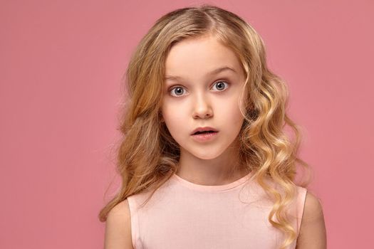 Beautiful little girl with a blond curly hair, in a pink dress is posing for the camera and looks wondered, on a pink background