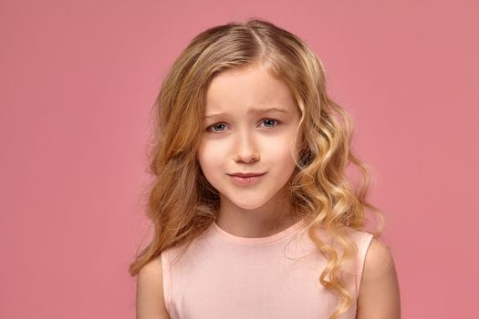 Nice little girl with a blond curly hair, in a pink dress poses for the camera and looks upset, on a pink background