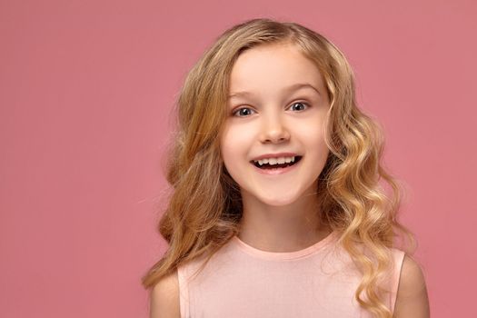 Gorgeous little girl with a blond curly hair, in a pink dress is smiling into the camera, on a pink background