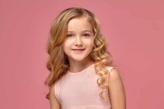 Sweet little girl with a blond curly hair, in a pink dress smiles for the camera and looks verry happy, on a pink background