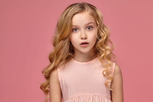 Charming little girl with a blond curly hair, in a pink dress poses for the camera and looks wondered, on a pink background