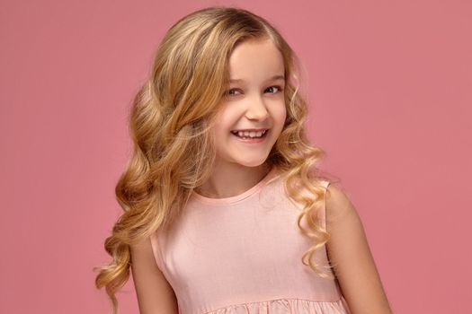 Little girl with a blond curly hair, in a pink dress is posing for the camera and smiling broadly, on a pink background