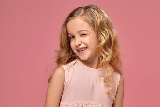 Little girl with a blond curly hair, in a pink dress winks and smiles into the camera, on a pink background