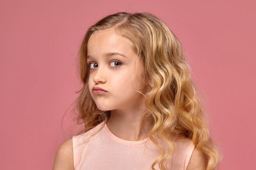 Beautiful little girl with a blond curly hair, in a pink dress poses for the camera and looks offended, on a pink background