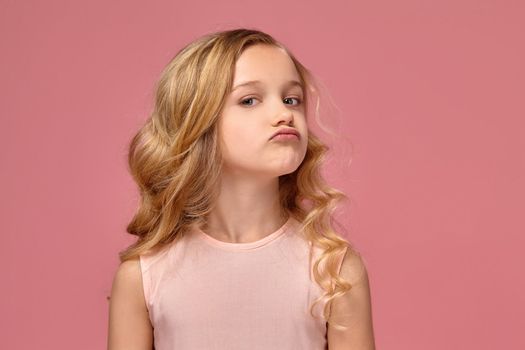 Pretty little girl with a blond curly hair, in a pink dress poses for the camera and looks offended, on a pink background