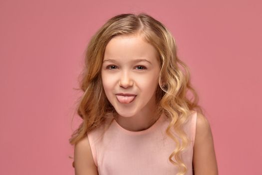 Beautiful little girl with a blond curly hair, in a pink dress poses for the camera and shows her tongue, on a pink background