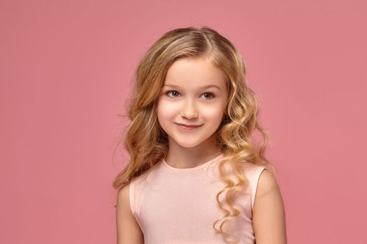 Amazing little girl with a blond curly hair, in a pink dress is smiling into the camera, on a pink background