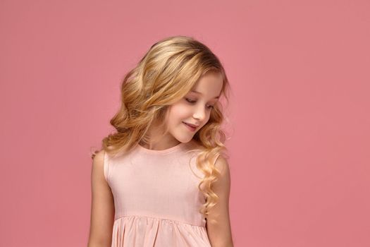 Beautiful little girl with a blond curly hair, in a pink dress poses for the camera and looks shy, on a pink background