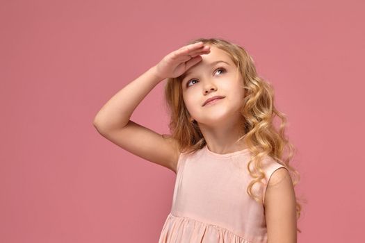 Beautiful little girl with a blond curly hair, in a pink dress is posing for the camera and looking up, on a pink background