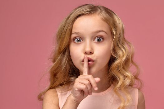 Lovely little girl with a blond curly hair, in a pink dress shows a gesture quiter, on a pink background