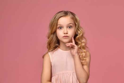 Sweet little girl with a blond curly hair, in a pink dress poses for the camera and looks wondered, on a pink background