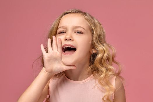 Beautiful little girl with a blond curly hair, in a pink dress is screaming at the camera, on a pink background