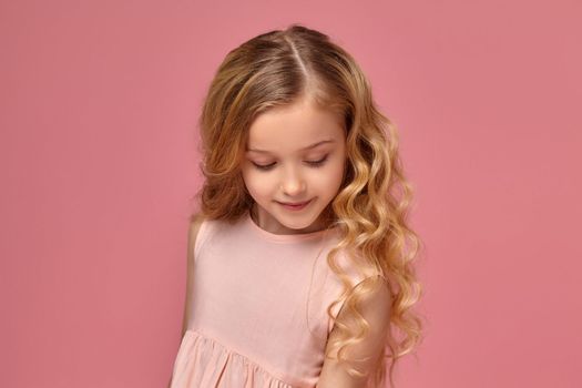 Pretty little girl with a blond curly hair, in a pink dress is posing fot the camera and looking shy, on a pink background