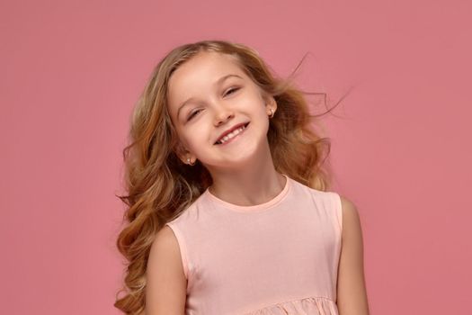 Funny little girl with a blond curly hair, in a pink dress is smiling into the camera, on a pink background