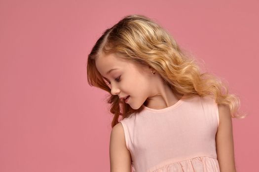 Gorgeous little girl with a blond curly hair, in a pink dress looks shy, on a pink background