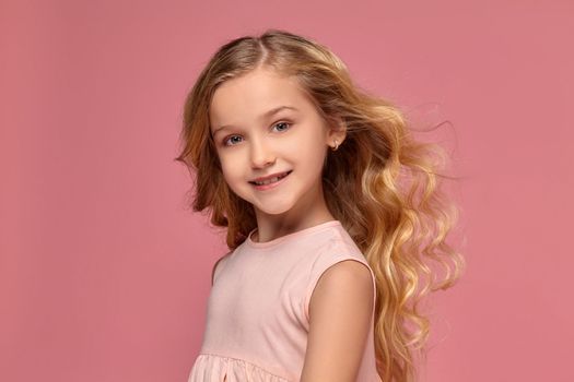 Lovely little girl with a blond curly hair, in a pink dress smiles for the camera and looks verry happy, on a pink background