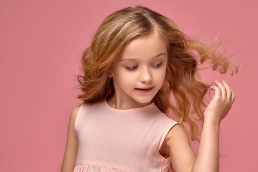 Little girl with a blond curly hair, in a pink dress poses for the camera and touches her hair, on a pink background