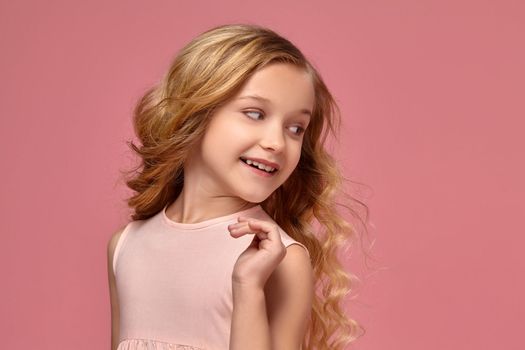 Beautiful little girl with a blond curly hair, in a pink dress is posing for the camera and looking back, on a pink background