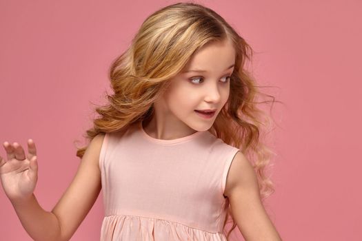 Funny little girl with a blond curly hair, in a pink dress is posing for the camera and looking away, on a pink background