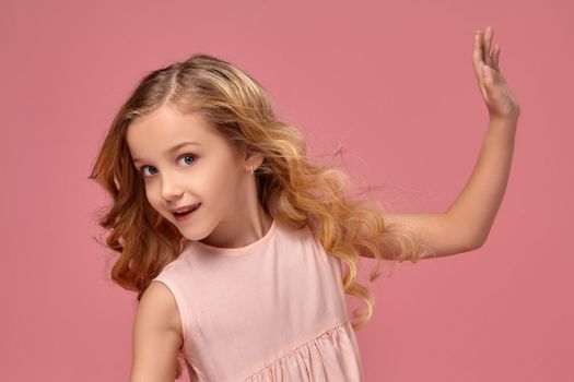 Beautiful little girl with a blond curly hair, in a pink dress poses for the camera and looks wondered, on a pink background