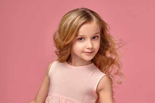 Beautiful little girl with a blond curly hair, in a pink dress is posing for the camera and looking wondered, on a pink background