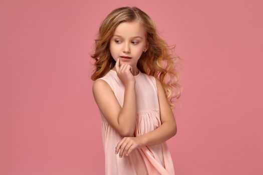Charming little girl with a blond curly hair, in a pink dress poses for the camera and looks thoughtful, on a pink background