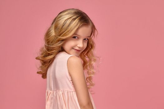 Beautiful little girl with a blond curly hair, in a pink dress is standing sideways and slyly smiling, on a pink background