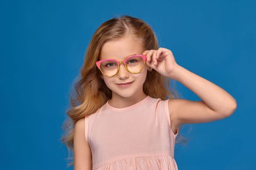 Modern little girl in a pink dress is straightense her fashionable glasses, standing on a blue background.