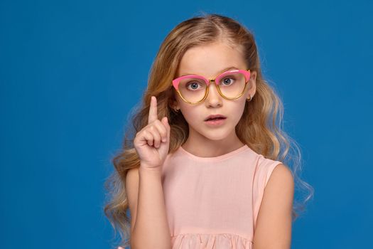 Pretty little girl in a pink dress and a fashionable glasses raised her finger up, on a blue background.