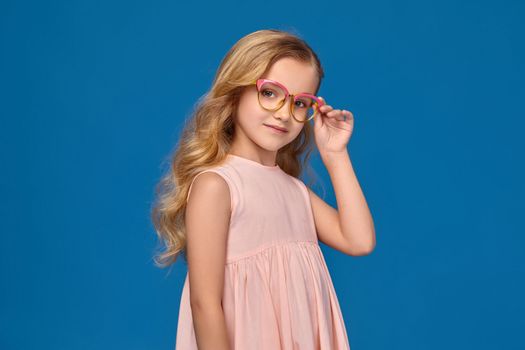 Beautiful little girl in a pink dress is straightense her fashionable glasses, standing on a blue background.