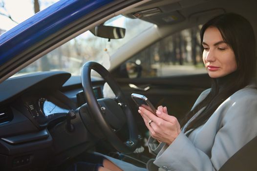 Confident successful middle-aged businesswoman, dressed in winter coat, sitting in the car on driver's seat, and using smart phone, making call to clients, planning business meetings. People Business