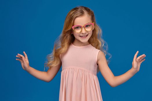 Modern little girl in a pink dress and a fashionable glasses is posing at the camera, standing on a blue background.