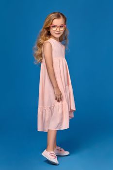 Modern little girl in a pink dress, fashionable glasses and a pink sneakers is looking at the camera, standing on a blue background.